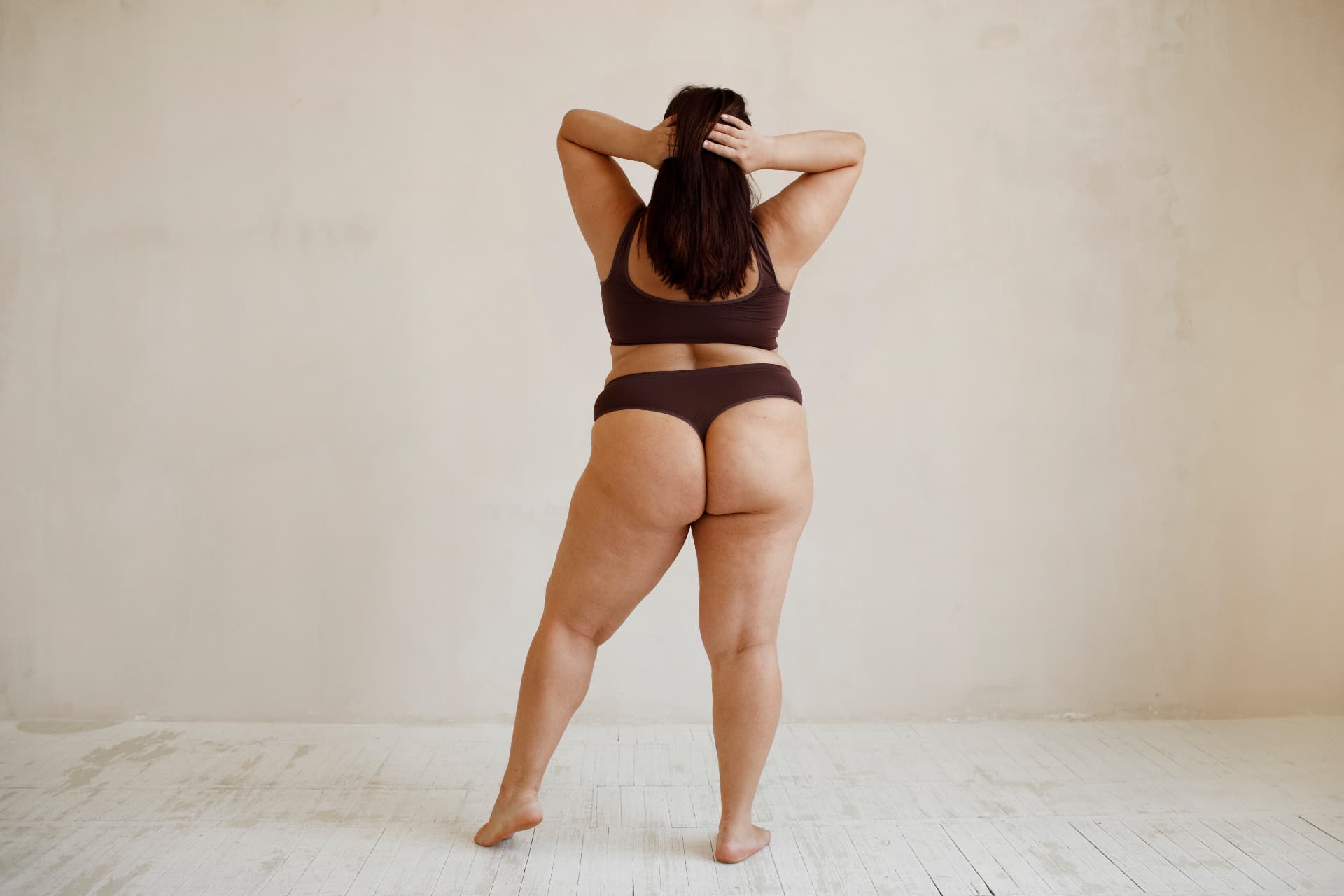 Woman standing confidently in brown compression lingerie, seen from behind in a sunlit studio